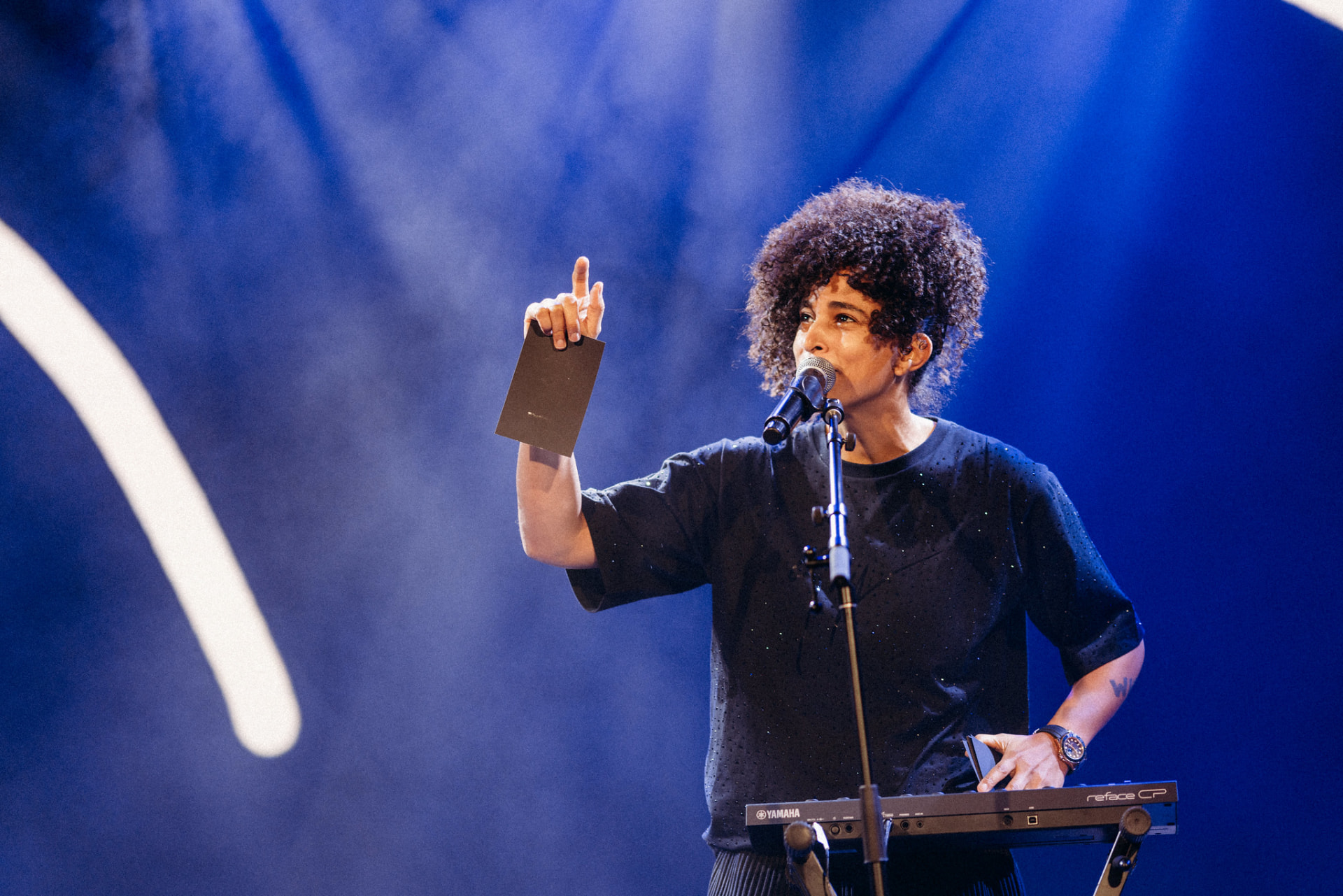Shantell Martin performing live on stage, holding up a black card at the microphone under blue stage lights