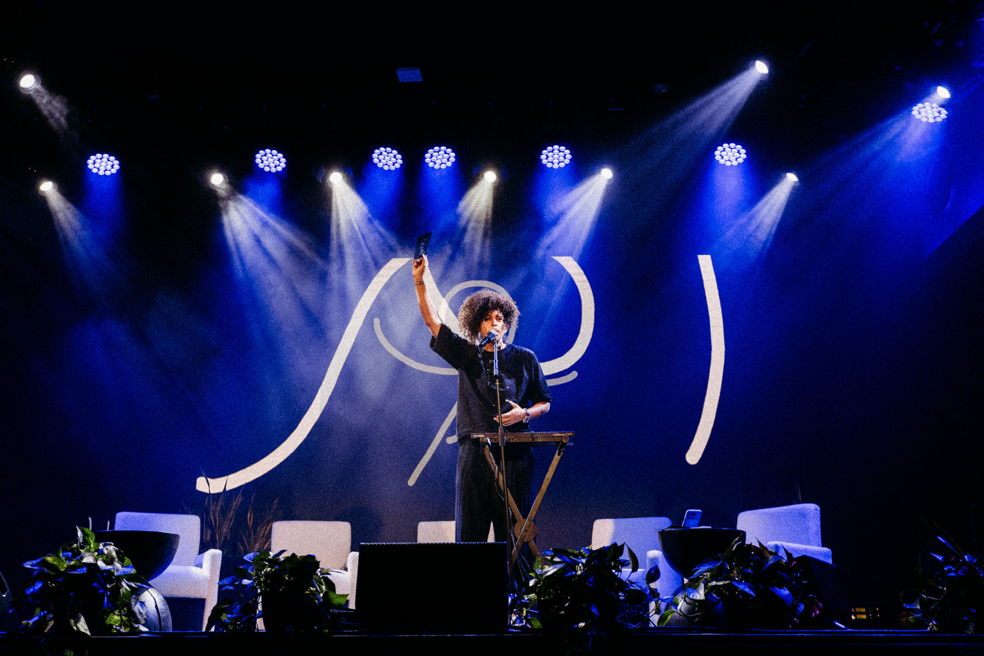 Shantell Martin performing live on a wide stage with plants, armchairs, and her signature white line projected behind her